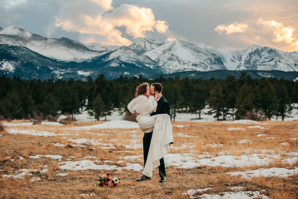 A groom holding his bride while kissing her. They are wearing wedding attire and standing in a field in Rocky Mountain National Park. The sun is setting over the mountains behind them.