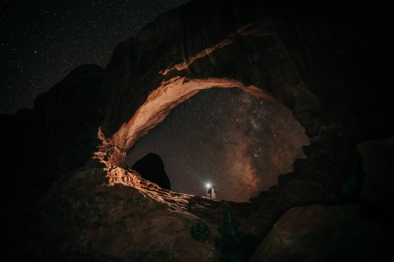Couple at night standing under a large eye-shaped arch during their Arches National Park elopement. The milky way is in the sky behind them.