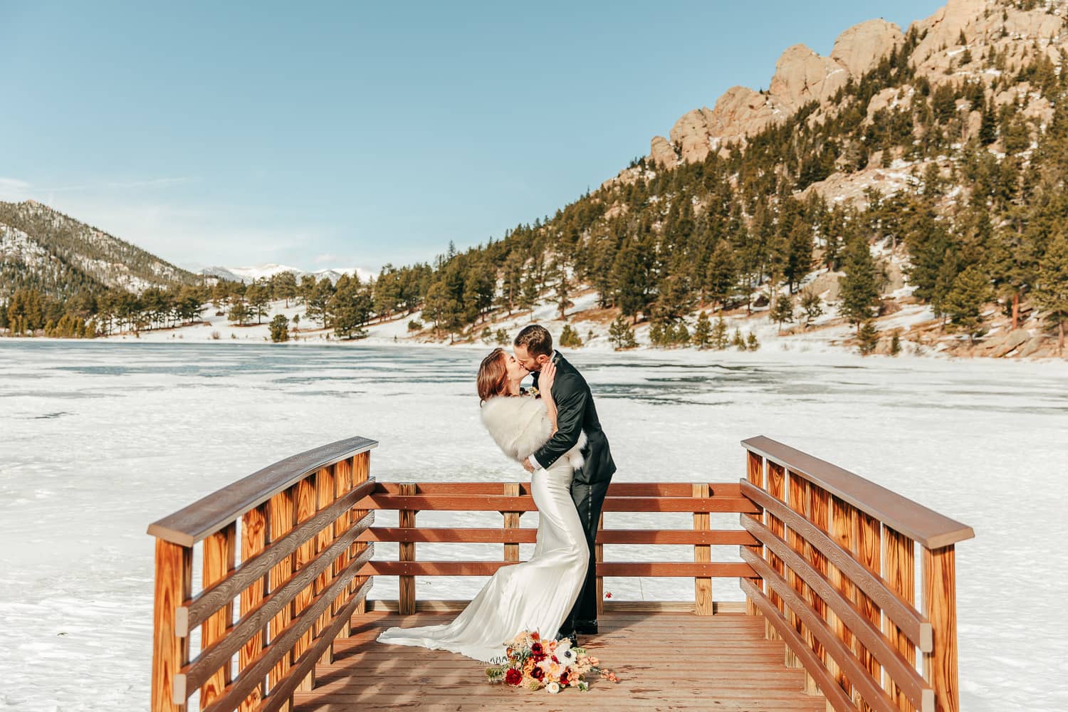 An elopement couple in wedding attire kissing on a lily lake dock in Rocky Mountain National Park.