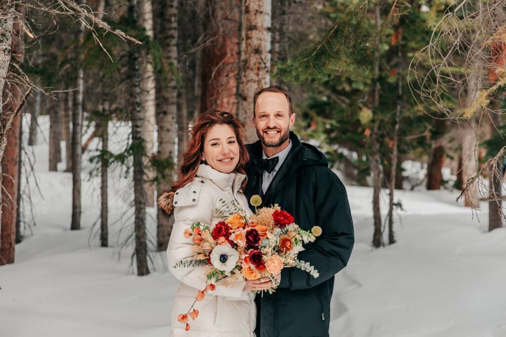 A couple in winter wedding attire. They are eloping in Rocky Mountain National Park. She is holding a colorful bouquet of flowers.