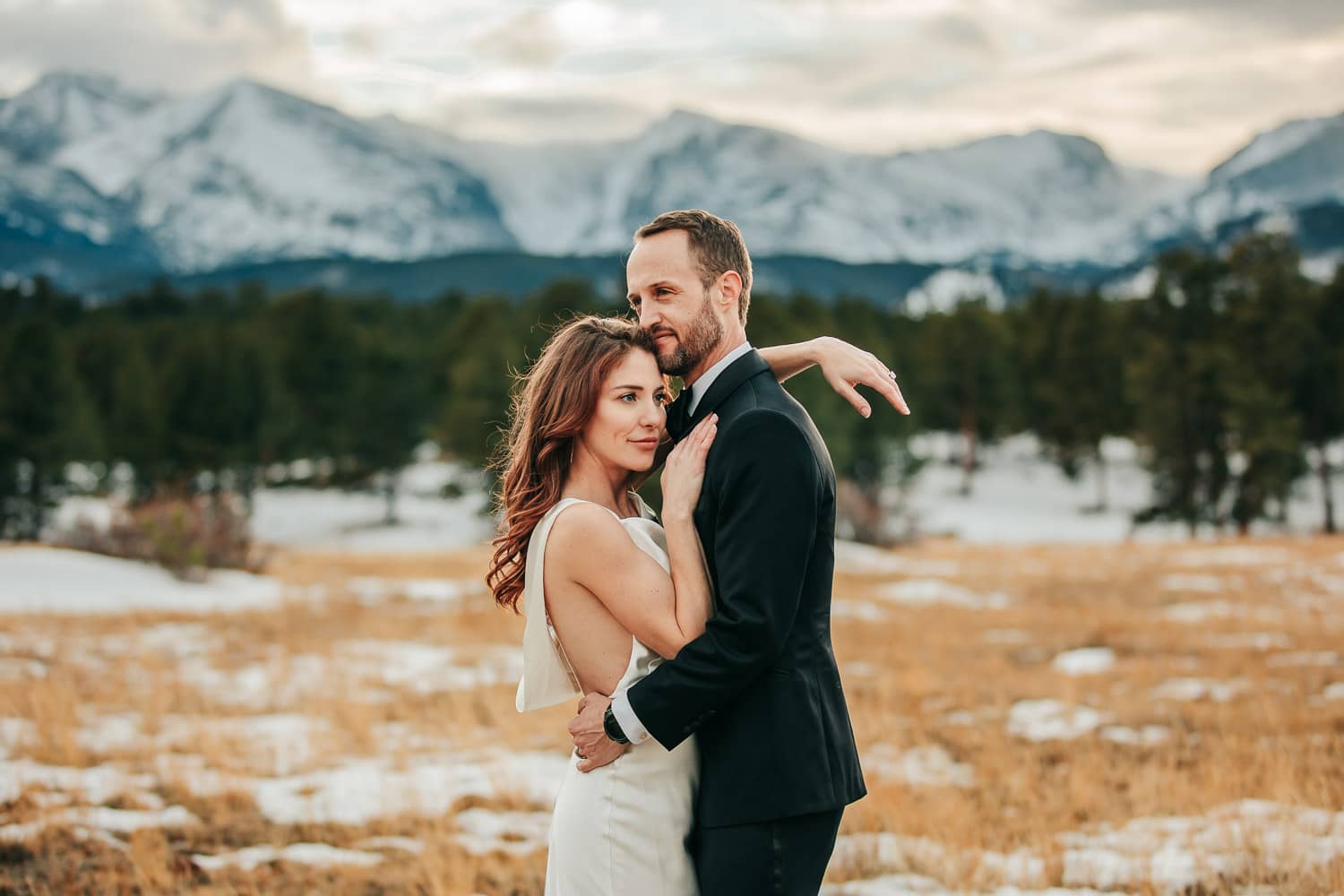 A couple in wedding attire standing close to each other, looking in opposite directions. The sun is setting over the colorado mountains behind them.