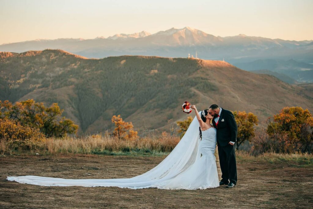 A couple in wedding attire during their colorado off-roading elopement. The bride has a very long veil on, and the couple is kissing while the bride lifts her bouquet in the air.