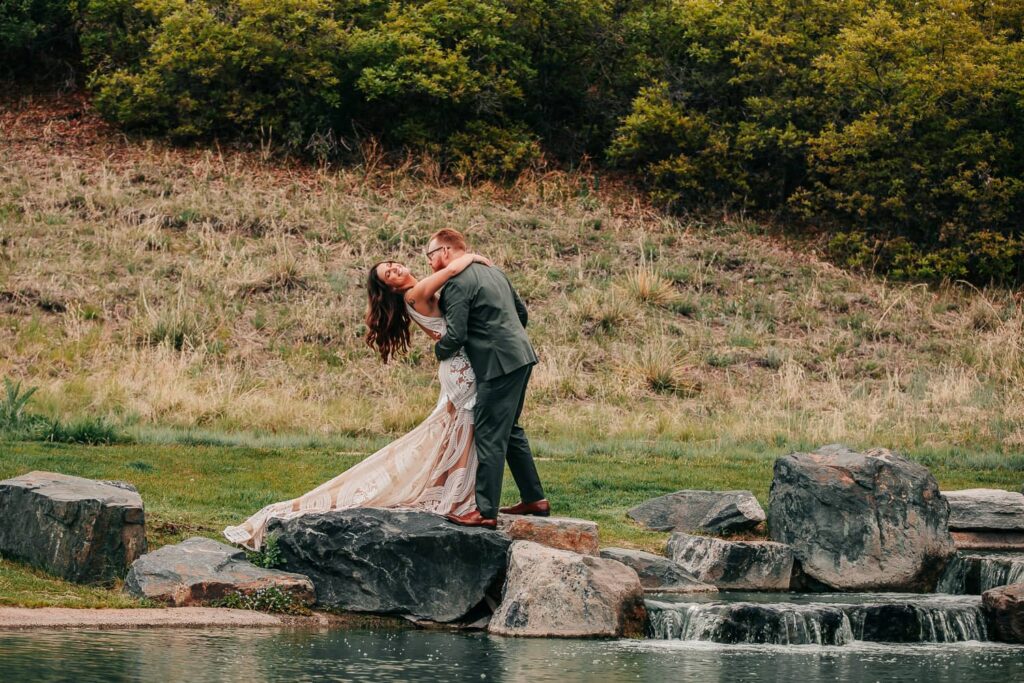 A couple in wedding attire during their colorado wedding. They are standing on the edge of a lake and waterfall while the groom dips the bride backwards and she laughs.