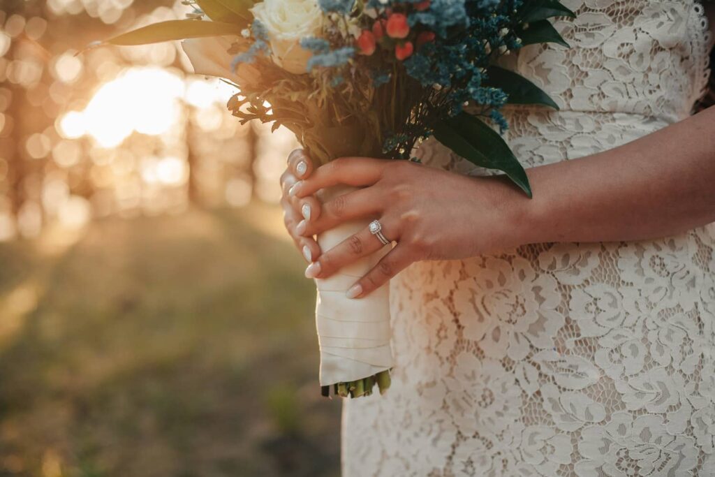 A bride during her colorado elopement. The photo is cropped in on her hand, holding her elopement bouquet. The emphasis is on her wedding rings, and the sun is bleeding through the trees in the background.