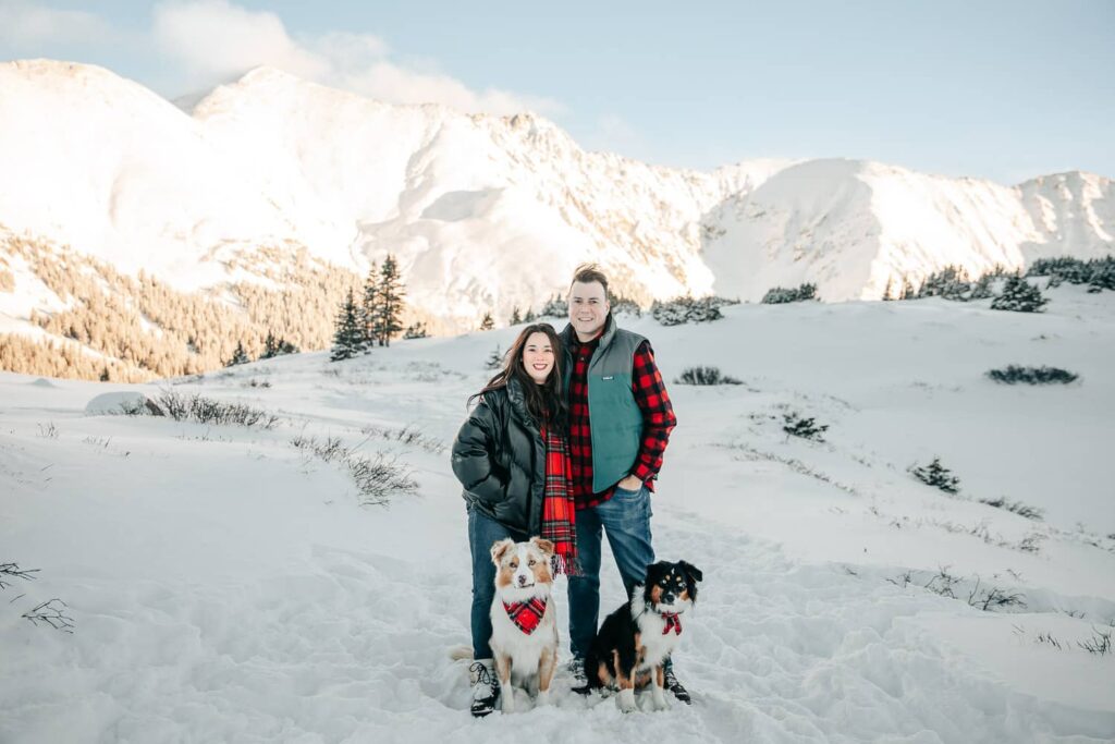 A couple and their dogs, wearing christmas style clothing. They are smiling at the camera while standing in the snow. There are big snow covered mountains in. the background.