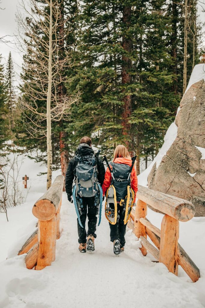 A couple hiking to their engagement photo location, they are crossing a wooden bridge and have large snowshoes on their hiking packs.