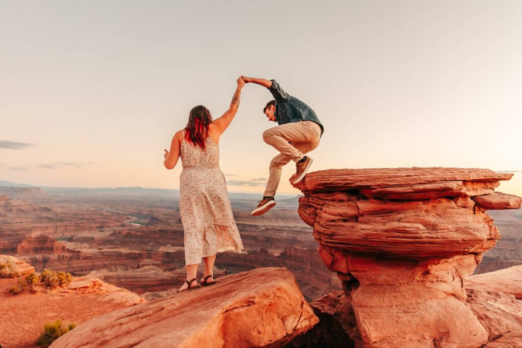 A couple in southern utah, the girl is holding her boyfriend's hand and he is jumping off of a rock.