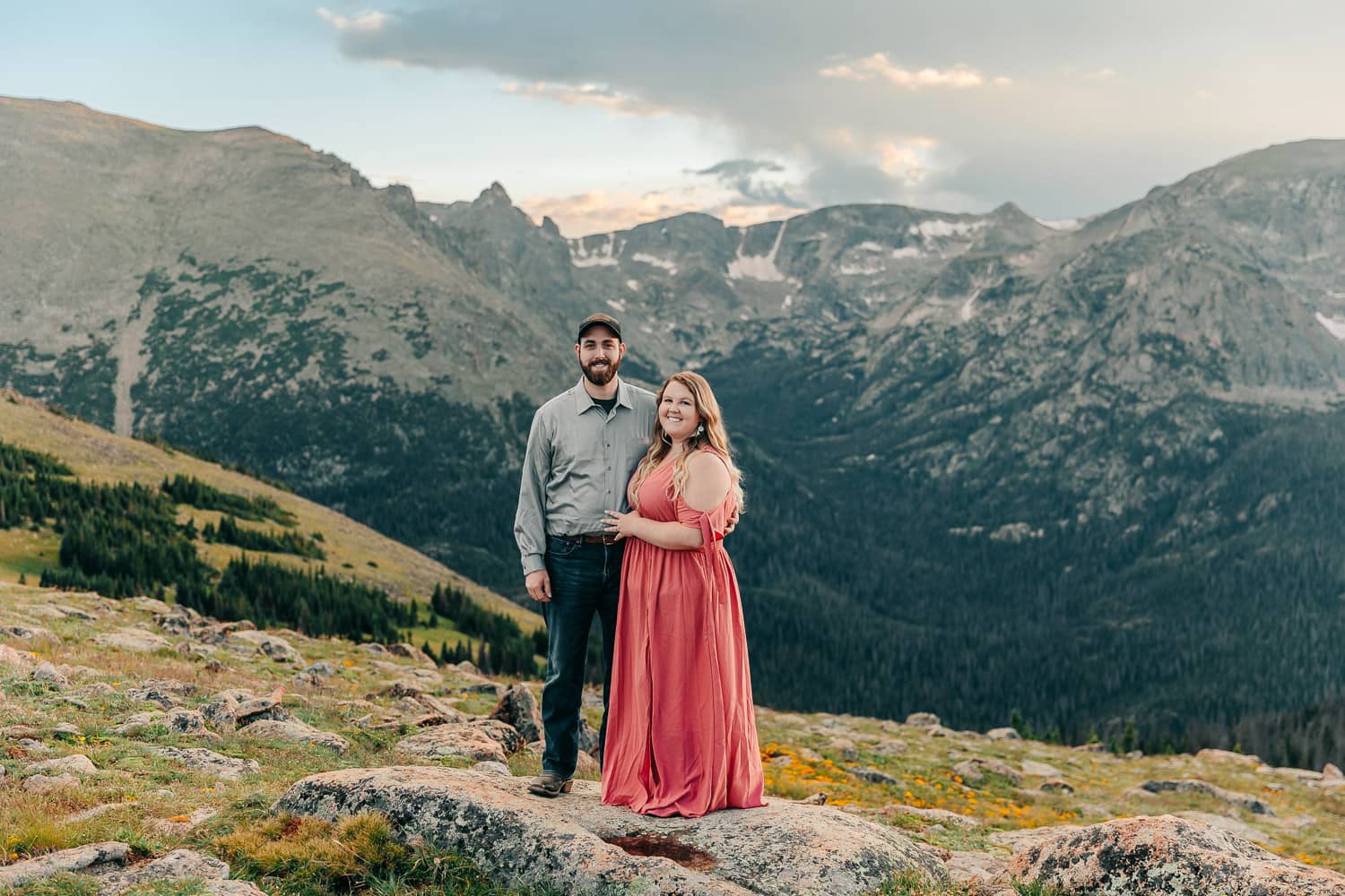 An engaged couple in formal attire, smiling. They are standing in front of a large mountain range at sunset in Rocky Mountain National Park Colorado.