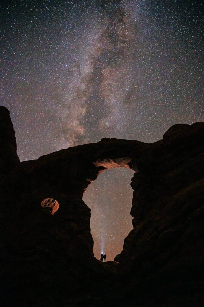 A couple standing in a large arch in Arches National Park at night. They are looking up with a headlamp. The milky way is visible behind them.
