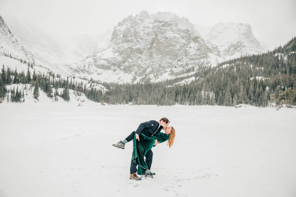 An engaged couple in formal wear on an alpine lake in front of a large mountain while it snows. He is dipping her back while kissing her.