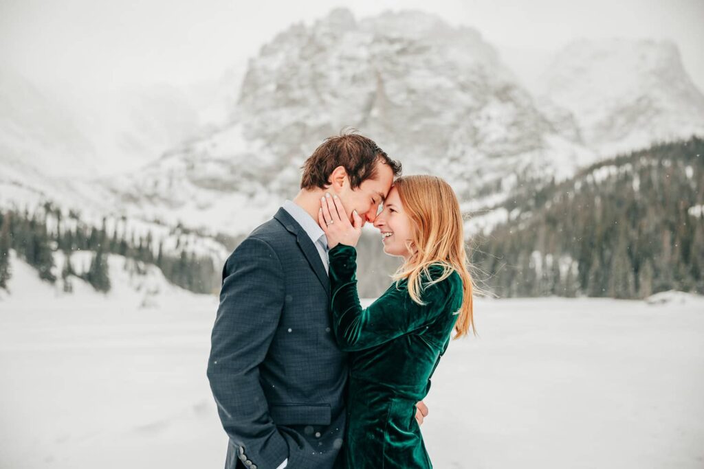 An engaged couple in formal wear leaning into each other in front of a large snowy mountain. They are standing on an alpine lake as it snows.