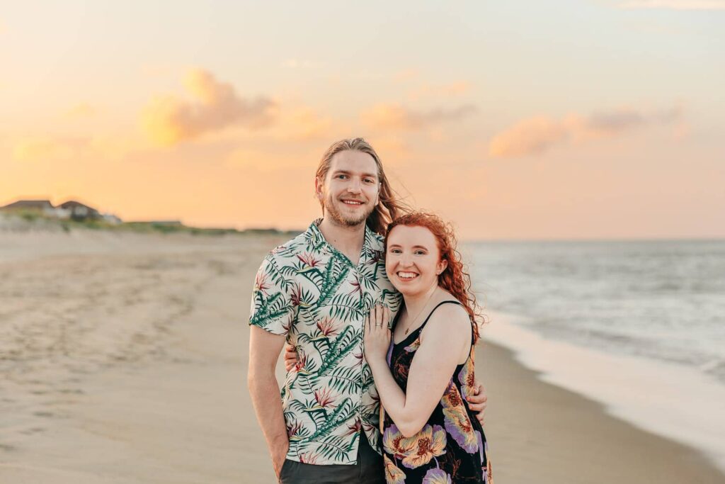 A couple smiling for a photo on a beach in North Carolina at sunset.