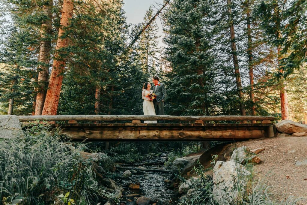 A couple in wedding attire standing on a wooden bridge above a creek. They are looking at each other. Rocky Mountain National Park elopement.