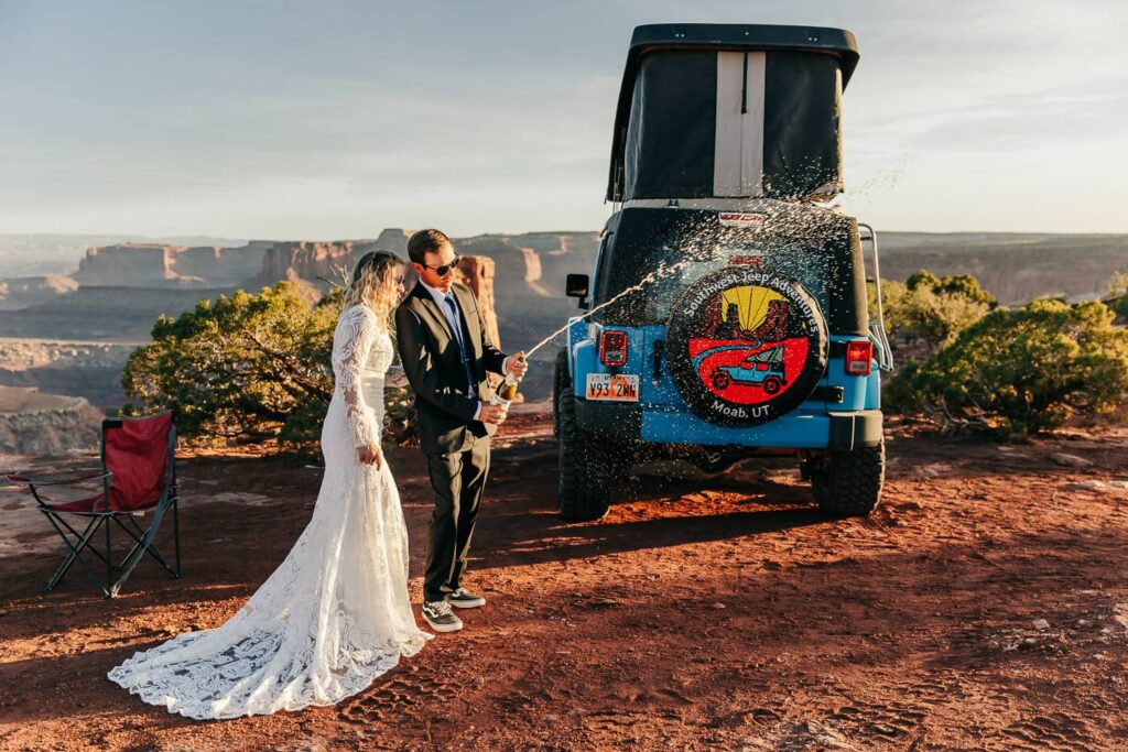 A couple in wedding attire popping champagne during their moab elopement. They are standing in front of a camper jeep.