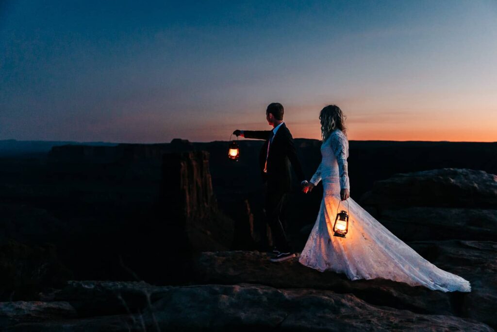 A wedding couple during their Moab elopement. They are holding lanterns and walking away together.