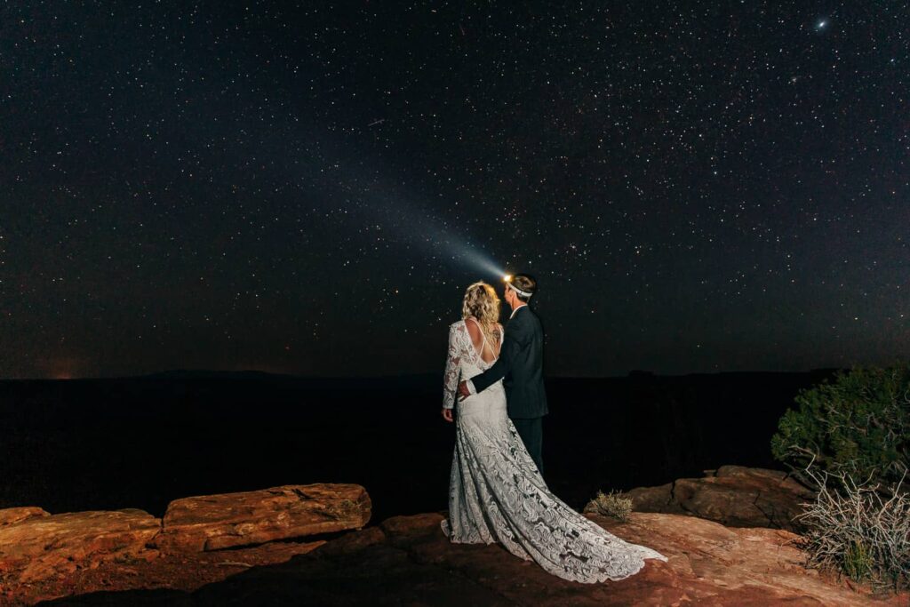 An eloping couple standing on a cliff in Moab, Utah at night. Looking away off into the stars.