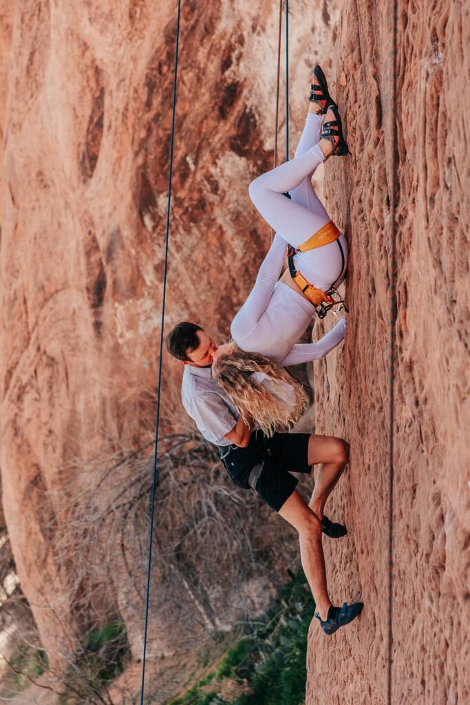 Climbing elopement in Moab, Utah. The bride is wearing all white and hanging upside down while her groom kisses her.