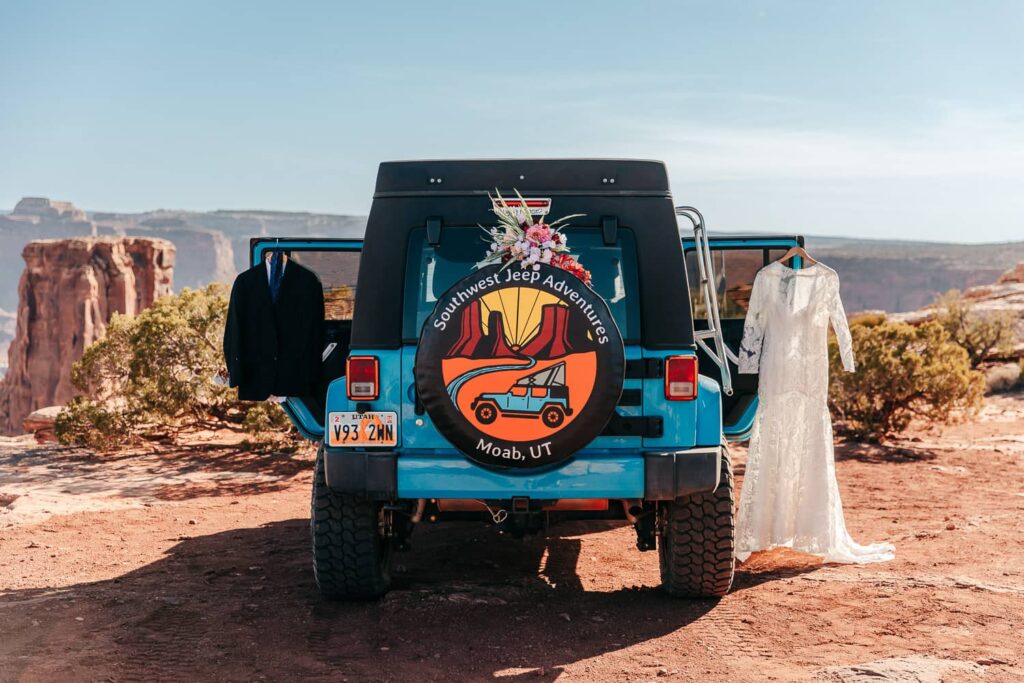 The back of a blue camper jeep with a wedding dress hanging on one door and a suit on the other. Bridal bouquet is on the spare tire. Getting ready for a moab elopement.