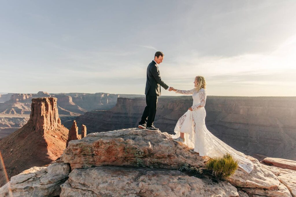 A groom helps his bride up a rock during their moab elopement. Canyons are in the background.