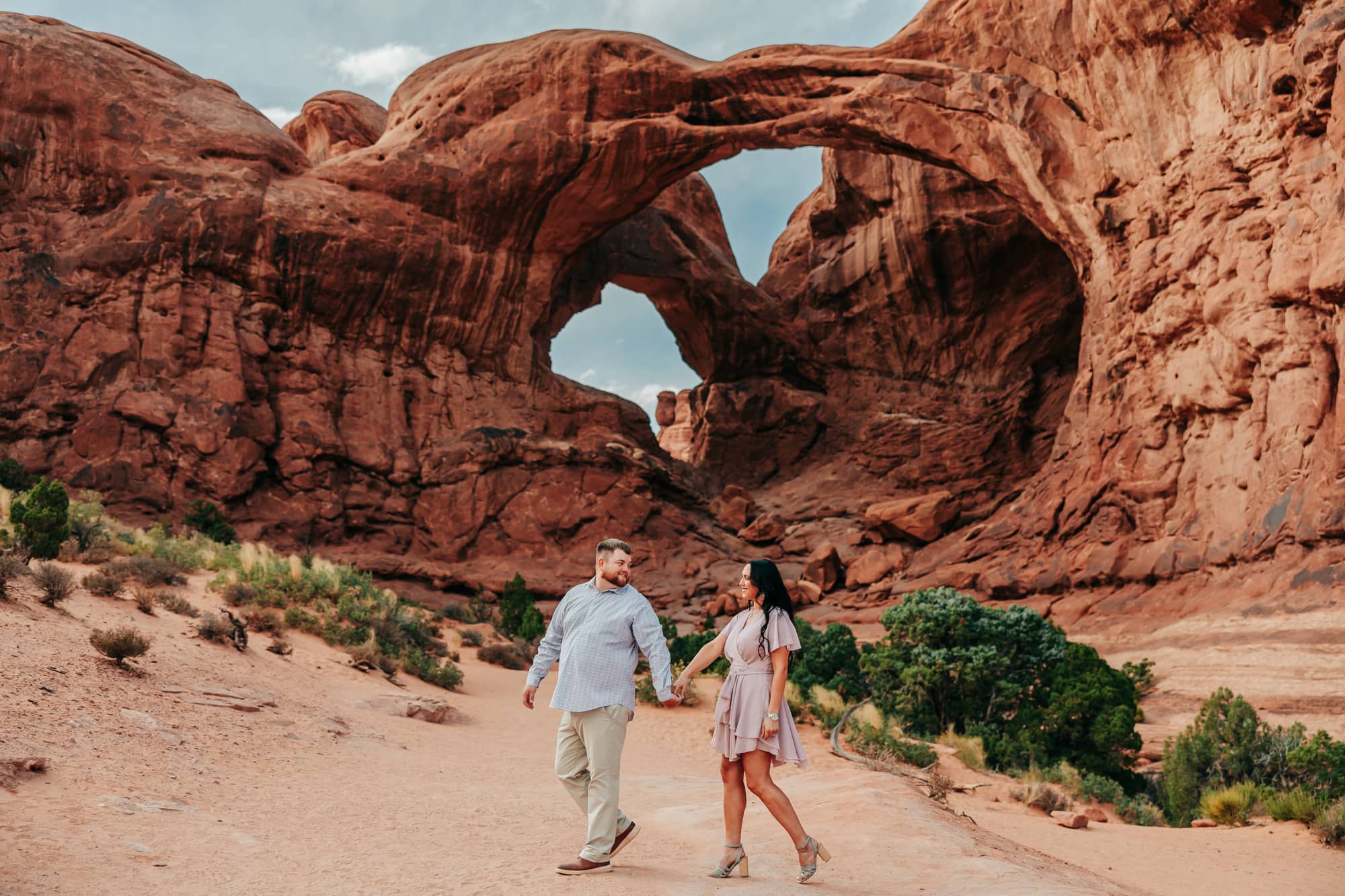 Couple holding hands and walking together in front of double arch in arches national park